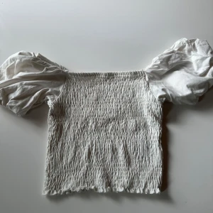 Vit smockad topp med puffärmar - Supersöt vit topp med smockad kropp och stora puffiga kortärmar. Offshoulder-modell som ger en trendig och romantisk vibe. Perfekt att styla med jeans eller kjol för en cool look.