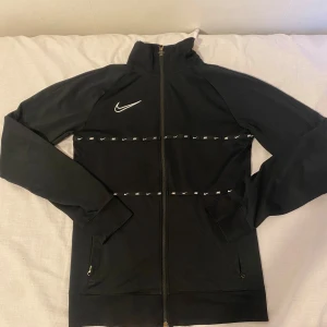 Svart Nike Dri-Fit vindjacka S - Svart Nike Dri-Fit vindjacka med hel dragkedja och höga krage. Jackan har vita Nike-loggor som detalj över bröstet och längs bröstet, samt två fickor med dragkedja. Perfekt för träning eller chill dagar.