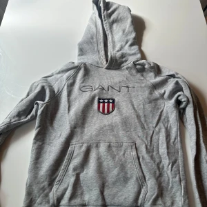 Grå hoodie från GANT med logga - Säljer en grå hoodie från GANT med broderad logga och sköld på bröstet. Tröjan har huva, magficka och långa ärmar. Perfekt för dig som gillar klassisk streetstyle och vill ha något bekvämt och snyggt. Passar dig som är ca 176 cm.
