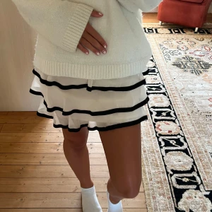 Ljusrosa/vit volangkjol med svarta ränder - Supersöt vit/ljusrosa kortkjol med volanger och svarta ränder. Kjolen har en luftig och flowy känsla, perfekt för att skapa en lekfull look. Materialet känns lätt och skönt mot huden, och de svarta detaljerna ger en snygg kontrast till det vita tyget.💛