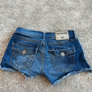 Blå jeansshorts True Religion - Snygga blå jeansshorts från True Religion med klassiska fickor bak och coola slitningar samt fransad kant. Shortsen har låg midja, knapp och dragkedja framtill samt detaljerade sömmar och logga på bakfickan. Perfekta för sommaren❤️