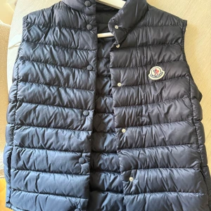 Mörkblå dunväst från Moncler - Snygg dunväst från Moncler. Västen har tryckknappar framtill och Moncler-logga på bröstet. Storlek 0 motsvarar Storlek Xs-S. Använd ett fåtal gånger och ser ny ut.