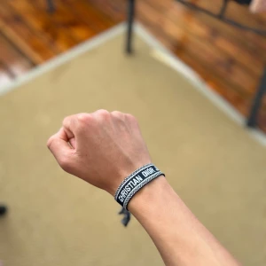 Christian Dior svart tygarmband - Svart och vitt tygarmband från Christian Dior med texten 'CHRISTIAN DIOR J'ADIOR' broderad runt om. Armbandet har en enkel och stilren design med knytning för justerbar passform. Perfekt accessoar för att lyfta din outfit med en lyxig touch.