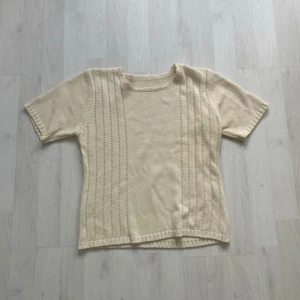 Beige stickad kortärmad tröja - Säljer en beige stickad tröja med korta ärmar och diskreta hålmönster i vertikala rader på framsidan. Tröjan har en rund hals med liten dekorativ kant och ribbad nederkant. Perfekt för dig som gillar enkel och stilren design.