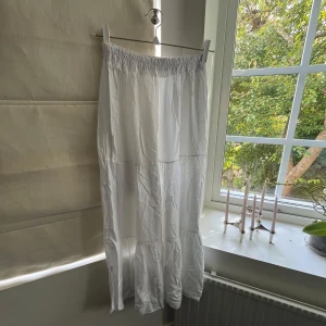 Vit långkjol med volang - Säljer en vit långkjol med elastisk midja och volangdetaljer. Kjolen är luftig och har en skön, flowy passform som gör den perfekt för varma dagar. Materialet känns som bomull och kjolen har en enkel, stilren look.