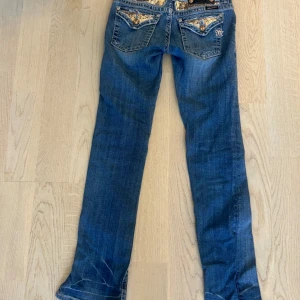 Miss Me blå jeans med detaljer, stl 26 - Snygga blå jeans från Miss Me med raka ben och coola broderade bakfickor med glittriga detaljer. Jeansen har klassisk femficksdesign, kontrastsömmar och är tillverkade i stretchig bomullsmix för skön passform. Dom har även en slits 