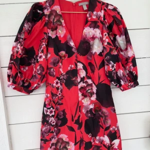 Röd, blommig klänning med puffärm - Röd kortklänning med stora puffärmar och djup v-ringning. Klänningen har ett färgstarkt blommigt mönster i rosa, svart och lila. Dragkedja i ryggen för enkel påklädning. 200kr + frakt