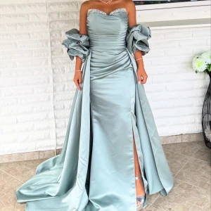 Ljusblå långklänning med puffärmar - Säljer en lyxig ljusblå långklänning med glittrande detaljer vid bysten och dramatiska puffärmar(avtagbara). Klänningen har en snygg slits framtill och är axelbandslös med en elegant omlott-effekt i midjan. Materialet är glansigt och ger en exklusiv känsla, perfekt för bal eller fest. 