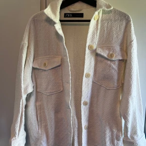 Vit jacka från Zara med struktur - Säljer en vit overshirt/jacka från Zara med snygg struktur i tyget och stora knappar framtill. Jackan har två bröstfickor med lock och knappar, samt klassisk krage. Perfekt att slänga över en hoodie eller t-shirt för en chill look.