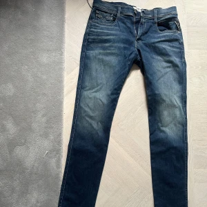 Replay Anbass Hyperflex jeans blå W32 L32 - Slim fit Replay Anbass Hyperflex jeans i mörkblå tvätt med snygga slitningar. Klassisk femficksmodell med dragkedjegylf och bruna detaljer på bakfickan. Stretchigt material för extra komfort och rörelsefrihet.