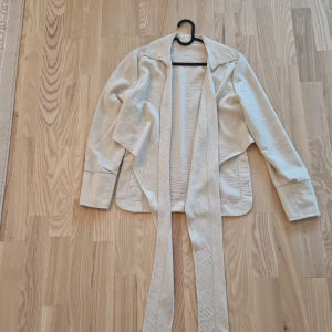 Beige omlott skjorta med bälte - Snygg beige skjorta med långärmad design och breda slag. Jackan har ett långt bälte som kan knytas omlott för en cool och avslappnad look. Linnématerialet är mjukt och har en lätt struktur, perfekt för lager-på-lager under vår och höst.