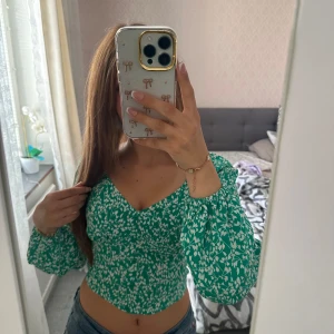 Grön blommig blus storlek XS - Supersöt grön blus med vitt blommönster från new yorker. Modellen är croppad med v-ringning och långa puffiga ärmar. Smockad rygg för extra bekväm passform. Perfekt för dig som vill ha en trendig och färgglad topp till våren eller sommaren! Använd en gång, skickar samma dag eller dagen efter💕