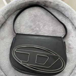 Svart axelväska med stor metallogo - Snygg svart axelväska i skinn med en bred axelrem. Diesel väska förutom att denna inte är äkta. Lite repor på själva loggan så inte värdens bästa skick. Skriv för mer bilder