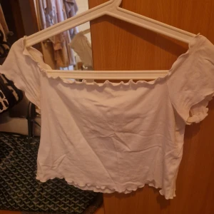 Vit offshoulder topp från Divided - Supersöt vit offshoulder topp från Divided med volangkant längs halsringning och nederkant. Croppad modell med korta ärmar och stretchigt bomullstyg. Perfekt till jeans eller kjol för en chill och trendig look.