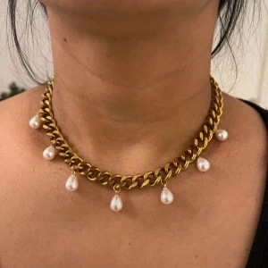 Guldhalsband med pärlhängen - Chunky guldhalsband med droppformade vita pärlor som hänger längs kedjan. Halsbandet har en cool och trendig vibe, perfekt för att lyfta din outfit med lite extra bling. Kedjan är grov och ger ett statement-utseende.