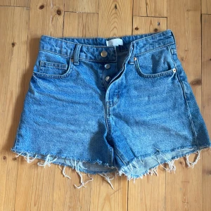 Blå jeansshorts från H&M, strl 34 - Snygga blå jeansshorts från H&M i klassisk femficksmodell med hög midja och råa, fransiga kanter. Shortsen har knappgylf och bälteshällor, perfekt för en chill sommarlook. Materialet är slitstarkt denim och färgen är ljusblå. Passar även storlek 36. Knappt använda och i nyskick❤️