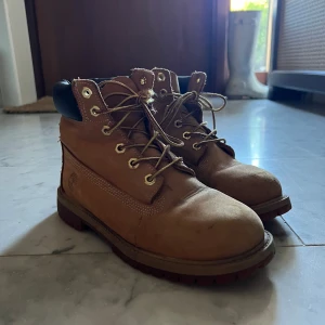 Bruna Timberland boots i skinn - Säljer ett par klassiska bruna Timberland boots i äkta skinn med robust snörning och guldfärgade öljetter. De har en vadderad svart krage och grov platt sula som ger grym komfort och stil. Perfekta för dig som gillar street och outdoor-vibe. De är använda och snörena har slitage men fyller fortfarande sin funktion. Viss slitage på lädret också som syns i bild.