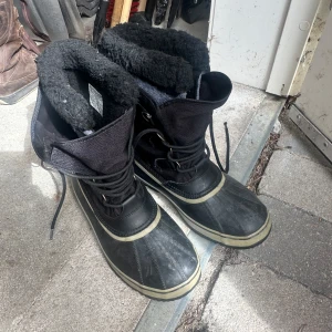 Svarta vinterkängor från Sorel - Rejäla svarta vinterkängor från Sorel med snörning och varm, mjuk fuskpäls på insidan. Yttermaterialet är en mix av textil och gummi, perfekt för kalla och blöta dagar. Klassisk design med rund tå och platt sula.