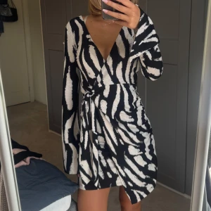 Zebramönstrad omlottklänning svart/vit - Snygg kort omlottklänning med zebramönster i svart och vitt. Djup v-ringning, långa ärmar och knyt i midjan för en smickrande passform. 
