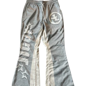 Gråa mjukisbyxor med tryck och snörning - Gråa mjukisbyxor med vita detaljer och breda ben. Byxorna har tryck längs ena benet, stjärndetaljer och texten 'Los Angeles only' på höften. Resår och snörning i midjan för skön passform. Perfekta för en chill och avslappnad stil.