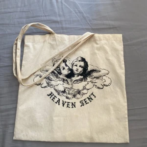 Beige shoppingväska från Brandy Melville - Säljer en beige shoppingväska ifrån Brandy Melville i bomull med svart tryck av en ängel bland moln och texten 'HEAVEN SENT' på framsidan. Väskan har långa axelband och är perfekt för att bära böcker, laptop eller snacks. Enkel och stilren design som passar till många outfits.