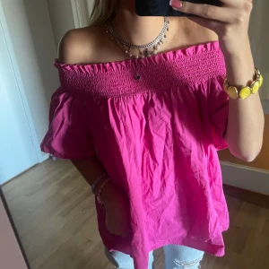 Offshoulder rosa blus med smock - Supersöt rosa offshoulder blus med smock upptill och puffiga korta ärmar. Luftig och flowy modell i bomull, perfekt för varma dagar. Snygg att styla med jeans eller kjol för en färgstark look.
