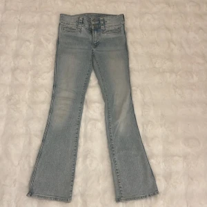 Ljusblå bootcut jeans med dubbla knappar - Snygga ljusblå jeans med bootcut passform och dubbla knappar i midjan. Jeansen har klassiska fickor fram och bak samt diskreta slitningar för en cool look. Perfekta för dig som gillar retrovibbar och vill ha ett par jeans som sticker ut.