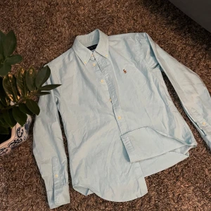 Ljusblå skjorta från Ralph Lauren - Klassisk ljusblå skjorta från Ralph Lauren med broderad logga på bröstet. Skjortan har lång ärm, knappar framtill och button down-krage. Tillverkad i bomull för en skön och stilren känsla. Perfekt för dig som gillar preppy och tidlös stil.