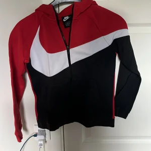 Nike hoodie röd svart vit med dragkedja - Nike hoodie i rött, svart och vitt med stor vit swoosh över bröstet. Tröjan har huva, hel dragkedja och känguruficka framtill. Långärmad och tillverkad i mjukt material, perfekt för chill eller träning. Snygg färgblockad design som sticker ut.