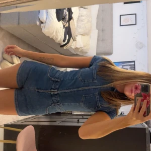 Jeansshorts jumpsuit med dragkedja - Snygg jeans jumpsuit i blå denim med korta ärmar, markerad midja och dragkedja framtill. Två stora bröstfickor och fickor bak ger en cool look. Perfekt för dig som gillar en avslappnad men trendig stil.