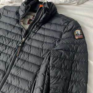 Svart dunjacka från Parajumpers XS - Snygg svart dunjacka från Parajumpers i storlek XS. Jackan har quiltad design, hög krage och dragkedja framtill. Märkets ikoniska patch sitter på ärmen och insidan är ljus med orange detaljer. Perfekt för kalla dagar och riktigt lätt att bära.