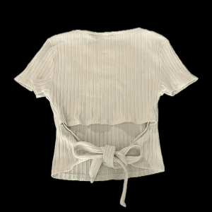 Vit ribbad topp med knyt i ryggen - Superfin vit ribbad topp från new Yorker med korta ärmar och rund halsringning. Toppen har en snygg knyt-detalj i ryggen som ger en unik look. Perfekt att styla med jeans eller kjol för en trendig outfit. Axelbredd-33cm. Längd-43cm.