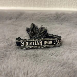 Christian Dior svart armband - Snyggt svart armband från Christian Dior med texten 'J'ADORE' broderad i vitt. Armbandet har dekorativa fransar i ändarna och ett stilrent, vävt mönster längs kanterna. Perfekt accessoar för att lyfta din outfit.