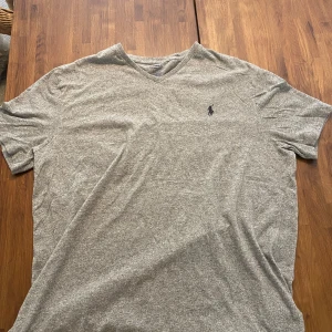 Grå v-ringad t-shirt från Polo Ralph Lauren - Grå t-shirt från Polo Ralph Lauren med v-ringning och klassisk logga broderad på bröstet. T-shirten är kortärmad och har en normal passform. Materialet är mjuk bomull som känns skönt mot huden. Perfekt för chill dagar och enkel att matcha med jeans eller shorts. Står L i tröjan men passar mig och jag har M 