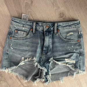 Blå jeansshorts med fransar - Säljer ett par blå jeansshorts från Mavi med råa, fransiga kanter och slitna detaljer. Klassisk femficksmodell med knapp och dragkedja framtill. Storlek står ej men skulle säga s