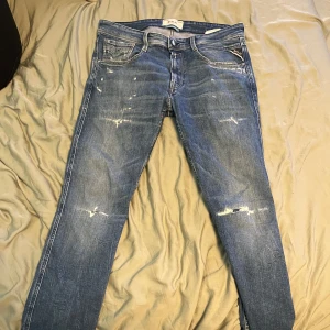 Replay jeans med slitningar - Säljer ett par blå jeans från Replay med snygga slitningar och tvättad look. Klassisk femficksmodell med raka ben och slim fit passform. Jeansen är tillverkade i bomull och har detaljerade sömmar på bakfickorna. Perfekta för en avslappnad streetstil. Storlek 31/32 bra skick! Bättre pris vid snabb affär 