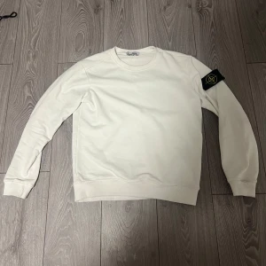 Vit sweatshirt från Stone Island - Säljer en stilren vit sweatshirt från Stone Island med klassisk rund hals och ribbade muddar. Tröjan har den ikoniska Stone Island-loggan på vänster ärm. Perfekt för dig som gillar enkel och clean streetwear. PASSFORM XS, FINNS ETT LITET FLÄCK I MITTEN. PRIS GÅR ATT DISKUTERA