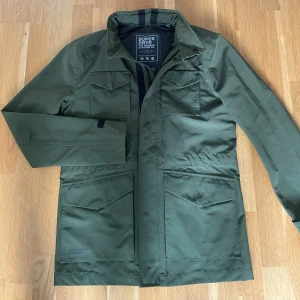 Olivgrön parkajacka från Superdry - Säljer en olivgrön parkajacka från Superdry med fyra stora fickor framtill och hög krage. Jackan har en clean och stilren look, perfekt för dig som gillar outdoor-vibe. Tillverkad i polyester och har en normal passform. Snygg detalj med Superdry-logga på ärmen. Känns som ny.