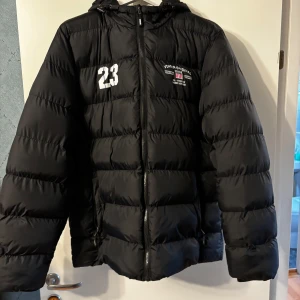 Svart pufferjacka från Vinson, L - Svart pufferjacka från Vinson med huva och dragkedja framtill. Jackan har tryck med siffran 23 och Vinson Original-logga på bröstet. Fylld för extra värme och har en sportig vibe. Perfekt för kalla dagar och enkel att matcha med streetwear.