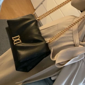 Svart axelväska från Dolce & Gabbana - Snygg svart axelväska från Dolce & Gabbana med guldfärgad kedja och logga framtill. Väskan har en mjuk, slät yta och stilren design som passar perfekt till din outfit. Kedjan ger en lyxig känsla och väskan är lagom stor för det viktigaste.