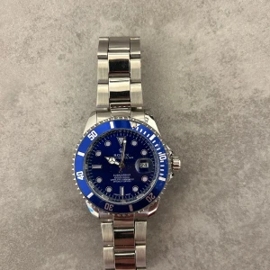 R Submariner blå urtavla - Snygg R Submariner med blå urtavla och blå vridbar bezel. Klockan har en robust länk i silverfärgat stål och tydliga indexmarkeringar. Klassisk rund form och datumfönster vid klockan tre. Perfekt för dig som gillar stilren design med sportig känsla.