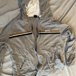 Grå kofta från BOSS - Snygg grå hoodie från BOSS med dragkedja, huva och fickor framtill. Tröjan har svarta, vita och bruna ränder över bröstet samt BOSS-logga. Storlek L. 
