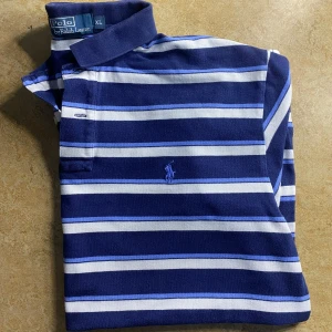 Randig pikétröja Polo Ralph Lauren XL - Klassisk pikétröja från Polo Ralph Lauren i blått, vitt och ljusblått randigt mönster. Tröjan har krage, knappar vid halsen och broderad logga på bröstet. Tillverkad i 100% bomull och passar perfekt till en avslappnad stil.