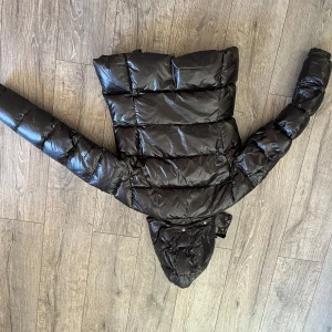 Svart pufferjacka med huva från wolff-wear  - Säljer en svart, glansig pufferjacka med avtagbar huva och dragkedja framtill. Jackan har långa ärmar, praktiska fickor med lock och tryckknapp på ärmen. Perfekt för kalla dagar och riktigt snygg streetstil.