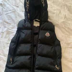 Svart dunväst från Moncler med huva - Säljer en svart dunväst från Moncler med stor huva och logga på bröstet. Västen har två dragkedjeförsedda fickor och Moncler-tryck på huvkanten. Perfekt för lager-på-lager och riktigt snygg till streetwear-stilen. Pris går att diskuteras, den passar bra som s
