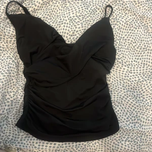 Svart omlott topp. Den har aldrig andvänts - Snygg svart topp med omlott-design och smala axelband. Modellen har en v-ringad skärning och lätt rynkade detaljer på sidorna för en cool och modern look. Perfekt för dig som vill ha en stilren och enkel topp med lite extra twist.
