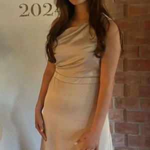 Champagnefärgad långklänning i satin - Säljer en elegant champagnefärgad långklänning med asymmetrisk nederdel och draperad halsringning. Klänningen har en mjuk satinfinish som ger ett lyxigt intryck och är ärmlös för en stilren look. Perfekt för speciella tillfällen!