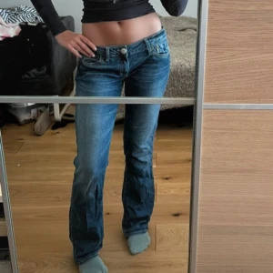 Blå bootcut jeans med låg midja - Snygga blå jeans med bootcut passform och låg midja. Klassisk femficksmodell med slitningar nertill på benen för en avslappnad vibe. Jeansen är i denim och har coola detaljer vid fickorna och knapparna.
