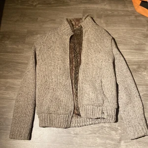 Beige stickad teddyjacka med fuskpäls - Mysig beige stickad teddyjacka med fuskpälsfoder på insidan. Jackan har ribbade muddar, hög krage och två fickor framtill. Perfekt för kalla dagar när du vill vara både varm och snygg. Stängs med dragkedja och har en mysig, avslappnad look. Finns även storleks reglering i midian. 