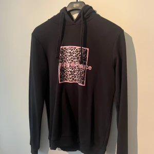 Svart hoodie med leopardprint från NA-KD - Svart hoodie från NA-KD i storlek XS med huva och snörning. Framsidan har ett rosa tryck med leopardmönster och texten 'strong & fierce'. Tröjan är långärmad och gjord i mjuk bomull, perfekt för chill dagar. Snyggt statement-plagg för dig som gillar djurmönster.